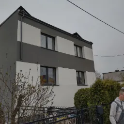 Dwukondygnacyjny dom z szaro-białą elewacją w poziome pasy, widoczny zza czarnego metalowego ogrodzenia i krzewów, pochmurne niebo w tle.