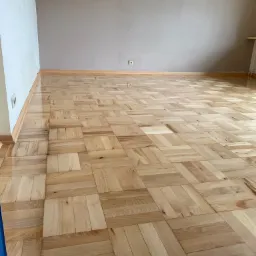 Parkiet ułożony w kwadraty, po cyklinowaniu, z widocznym gniazdkiem elektrycznym na ścianie w tle.