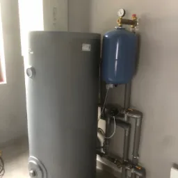 Szary, cylindryczny zbiornik buforowy ciepła z niebieskim naczyniem przeponowym i kompletem rur, zaworów oraz manometrem, zamontowany w pomieszczeniu z jasnymi ścianami i podłogą.