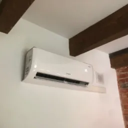 Biały klimatyzator marki Rotenso zamontowany na ścianie z widocznymi drewnianymi belkami stropowymi i fragmentem ściany z czerwonej cegły.