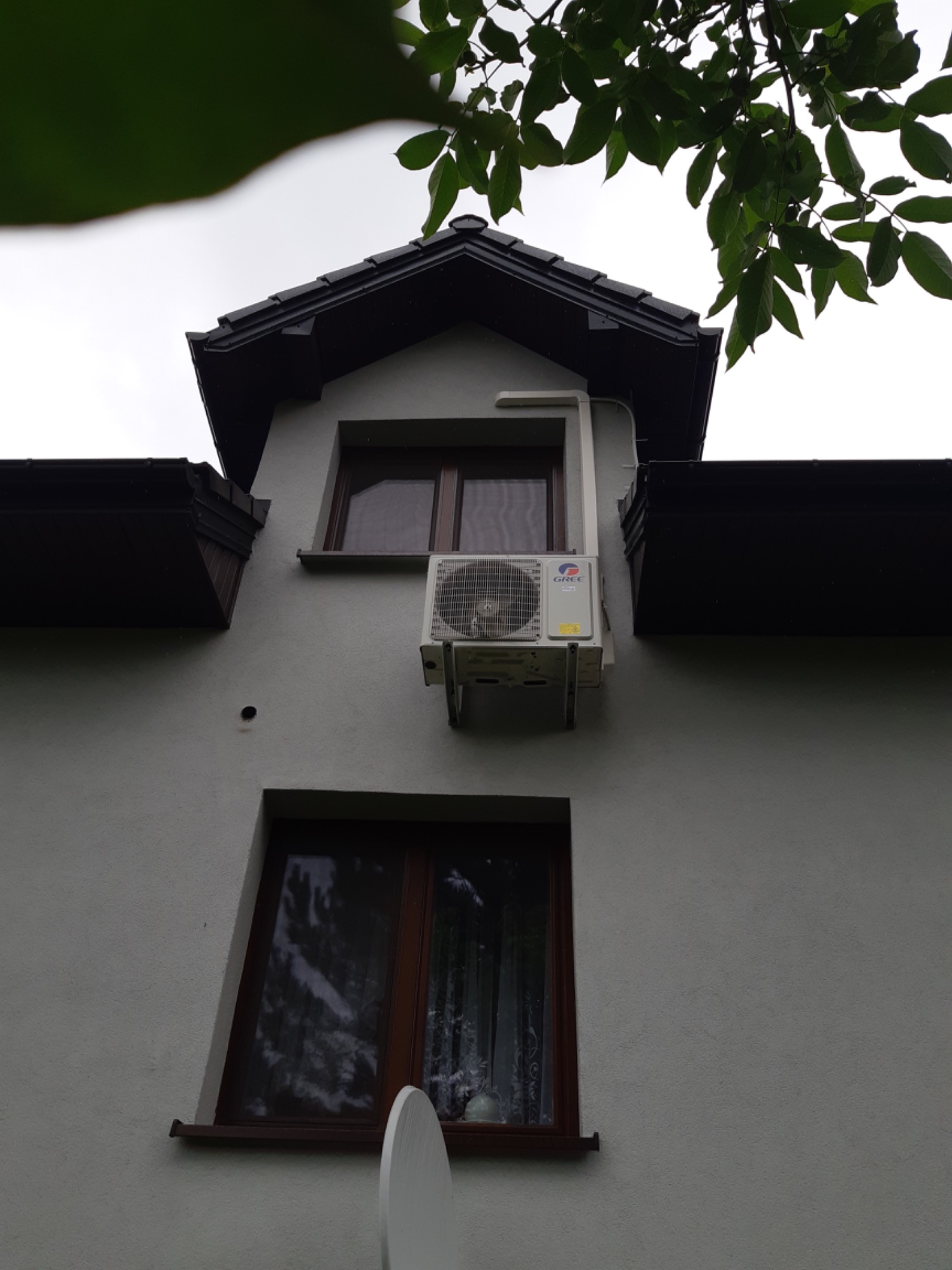 Biała jednostka zewnętrzna klimatyzacji marki Gree zamontowana na ścianie budynku, widok z dołu, fragment drzewa w górnej części kadru.