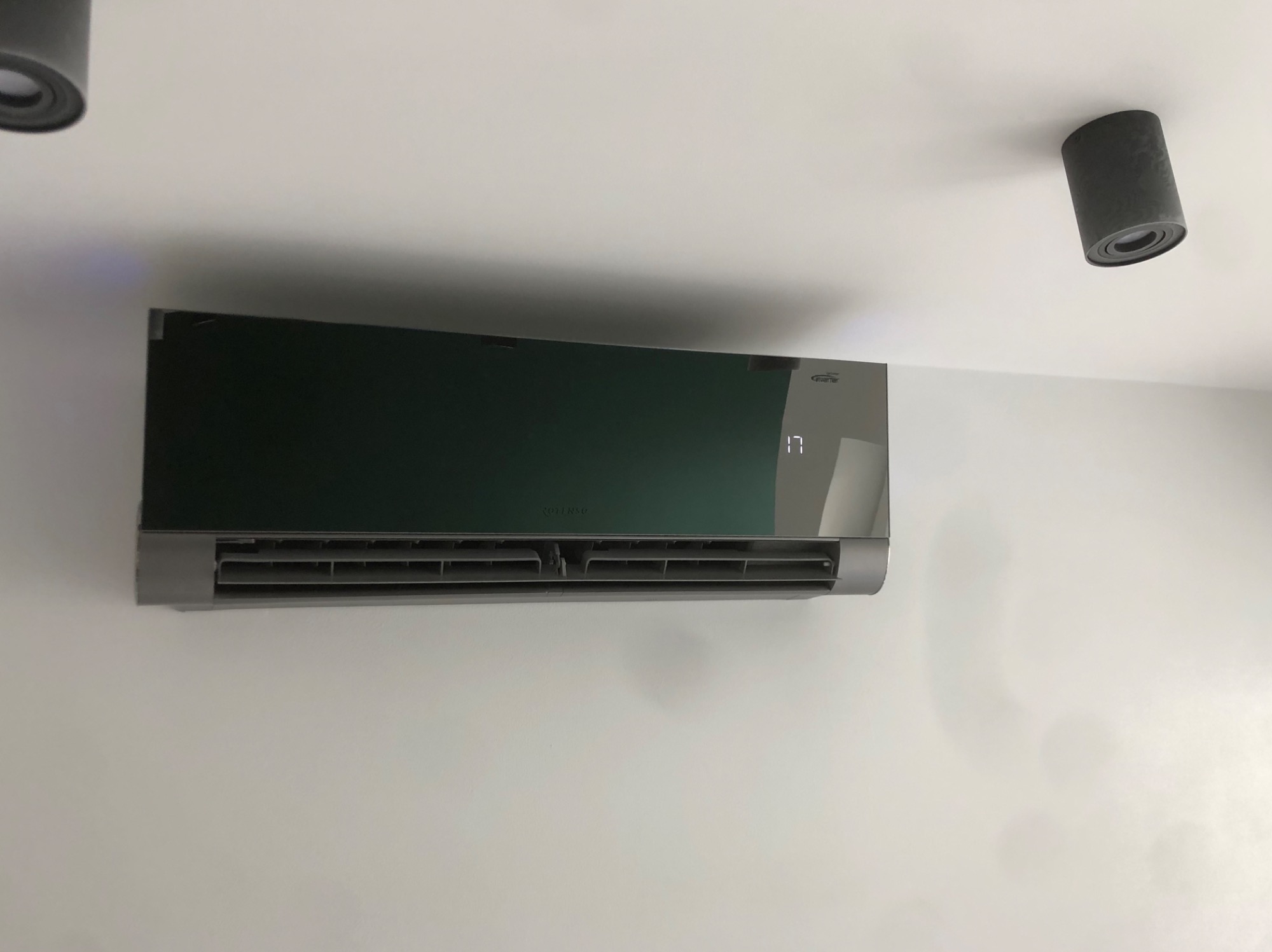 Nowoczesny klimatyzator ścienny w kolorze szaro-zielonym, zamontowany na białej ścianie, z widocznymi dwoma punktowymi lampami sufitowymi.