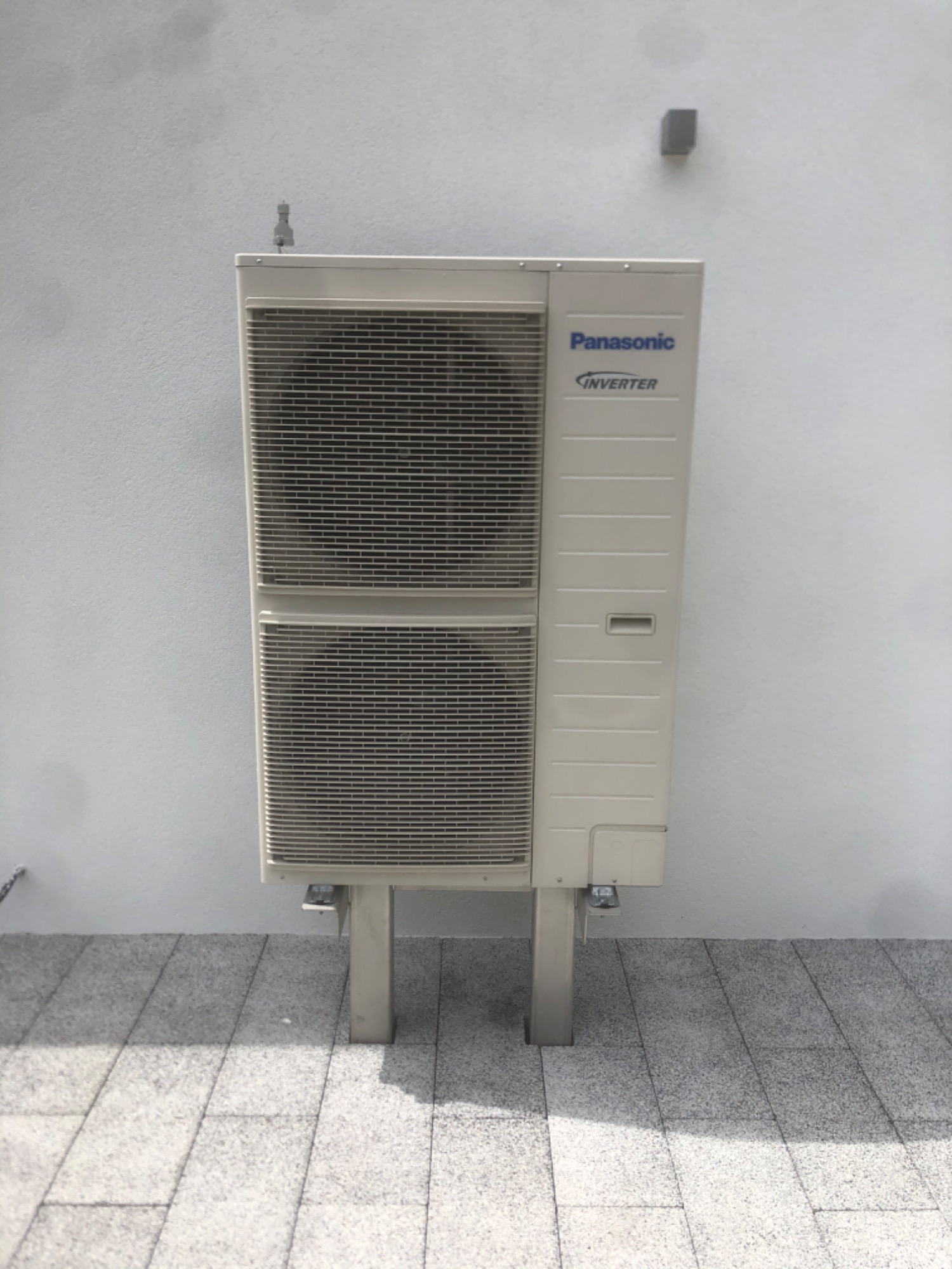 Zewnętrzna jednostka klimatyzacji Panasonic Inverter zamontowana na metalowych wspornikach przy jasnej ścianie, widoczna na tle szarej kostki brukowej.