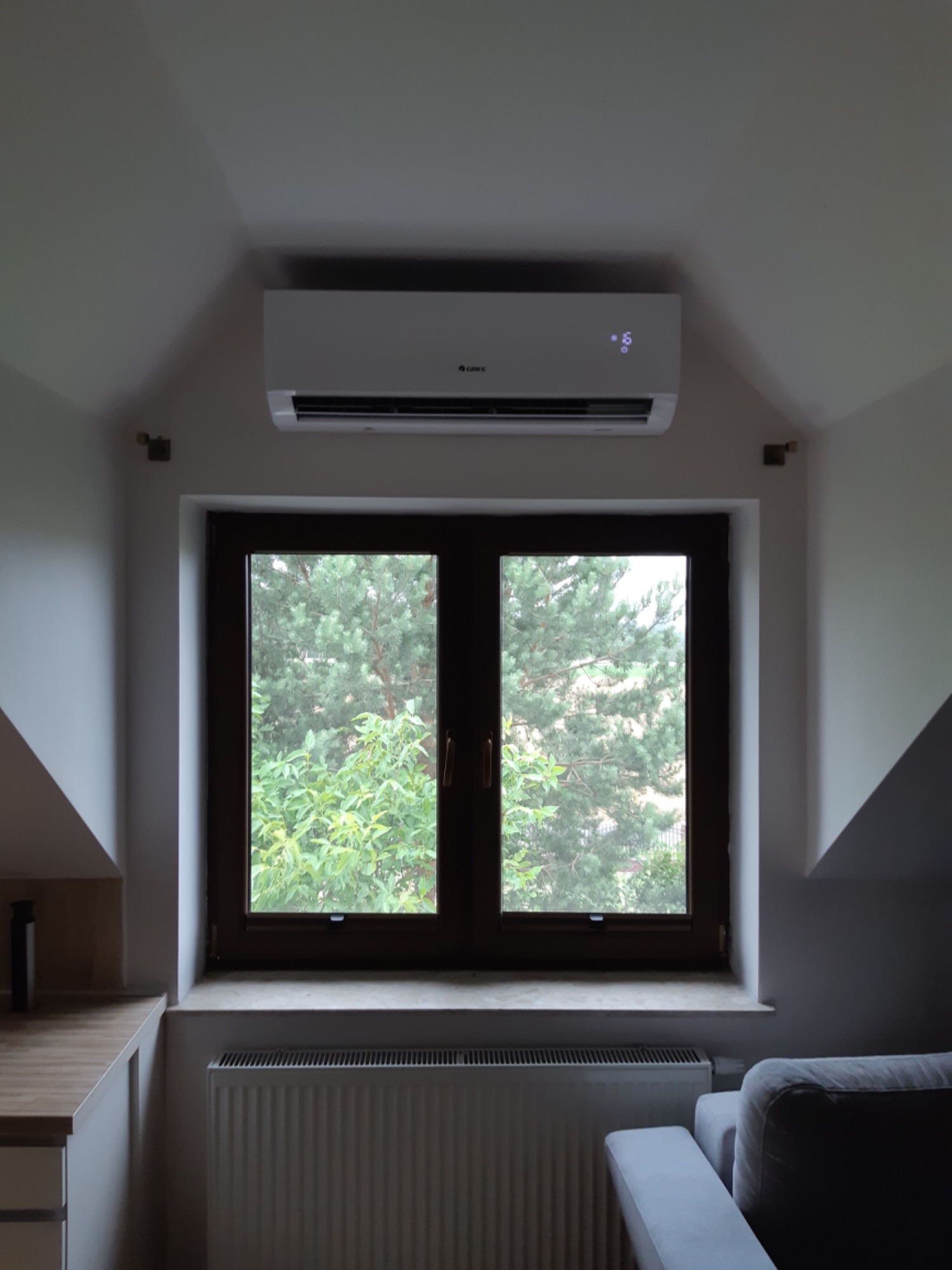 Biały klimatyzator marki Gree zamontowany nad oknem w pokoju na poddaszu, widoczne drzewa za oknem i grzejnik pod nim.