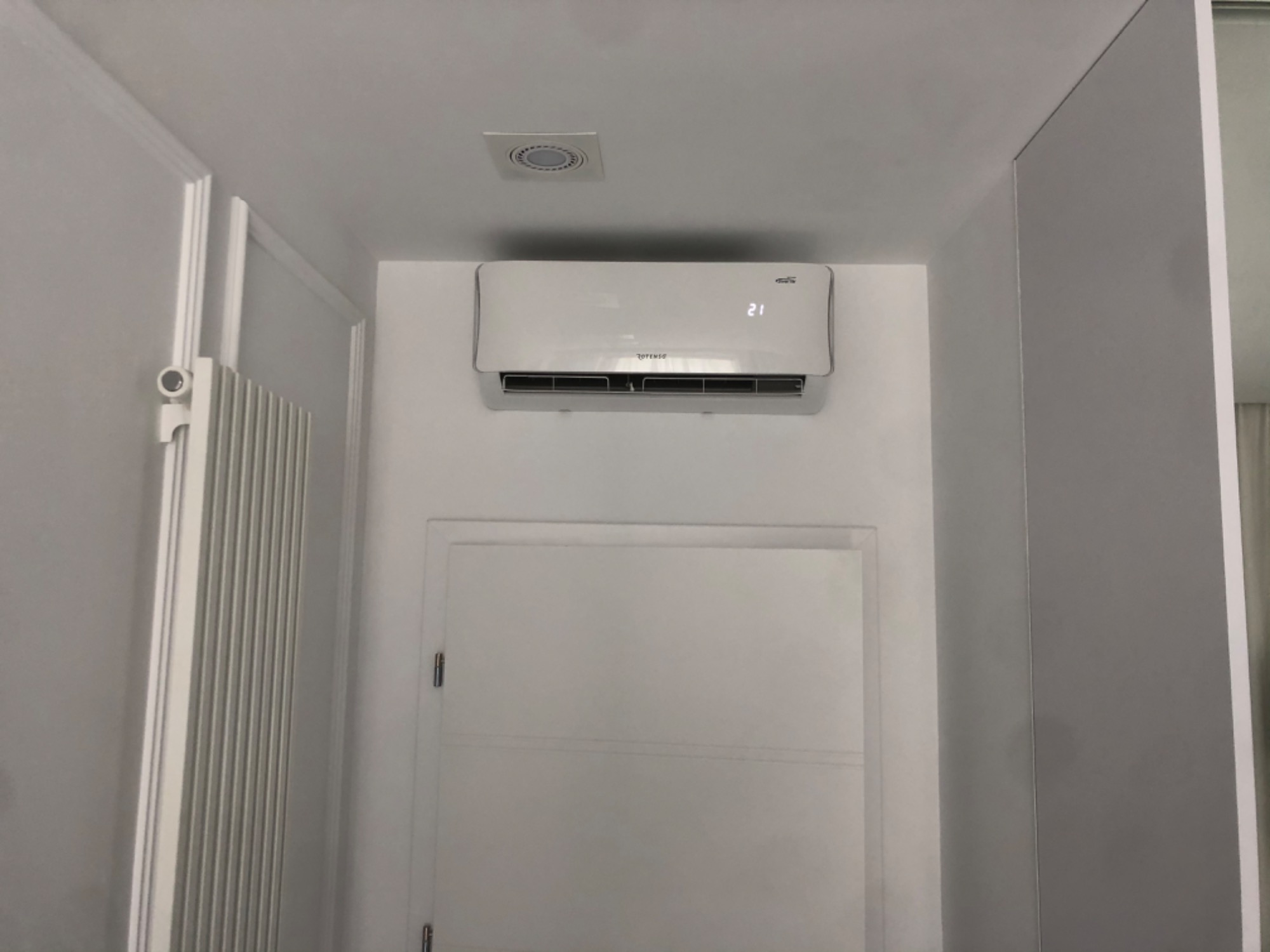 Biała jednostka klimatyzacji zamontowana na ścianie nad białymi drzwiami w minimalistycznym wnętrzu, widoczny fragment grzejnika i kratka wentylacyjna na suficie.