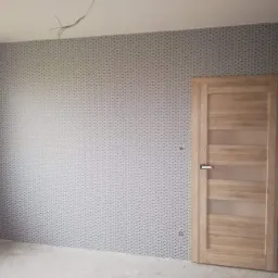 Pokój po tapetowaniu ścian wzorzystą tapetą w odcieniach szarości z drewnianymi drzwiami z przeszkleniami, widoczne wystające kable ze ściany.