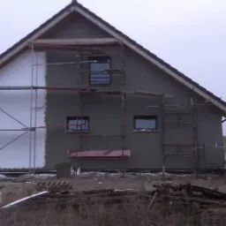 Dom w trakcie ocieplania: widoczne rusztowania, fragment ściany z izolacją styropianową, druga część pokryta szarym tynkiem. Wokół budynku porozrzucane materiały budowlane.