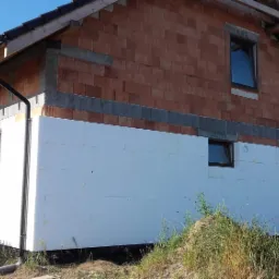 Dom w trakcie ocieplania: dolna część ściany pokryta białym styropianem, górna część z czerwonej cegły, widoczne okna i fragment dachu.