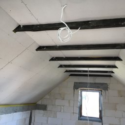 Krystian S. Docieplenia-remonty od A do Z - Poddasze w trakcie wykańczania: widoczne zamontowane płyty karton-gips na skośnym suficie z drewnianymi belkami, okno w surowej ścianie z białego bloczka, zwisający kabel elektryczny.