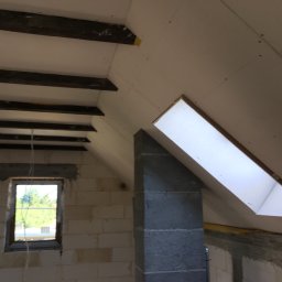 Krystian S. Docieplenia-remonty od A do Z - Poddasze w trakcie wykańczania: widoczne drewniane belki stropowe, okno dachowe, ściany z bloczków oraz zamontowane płyty kartonowo-gipsowe z widocznymi łączeniami.