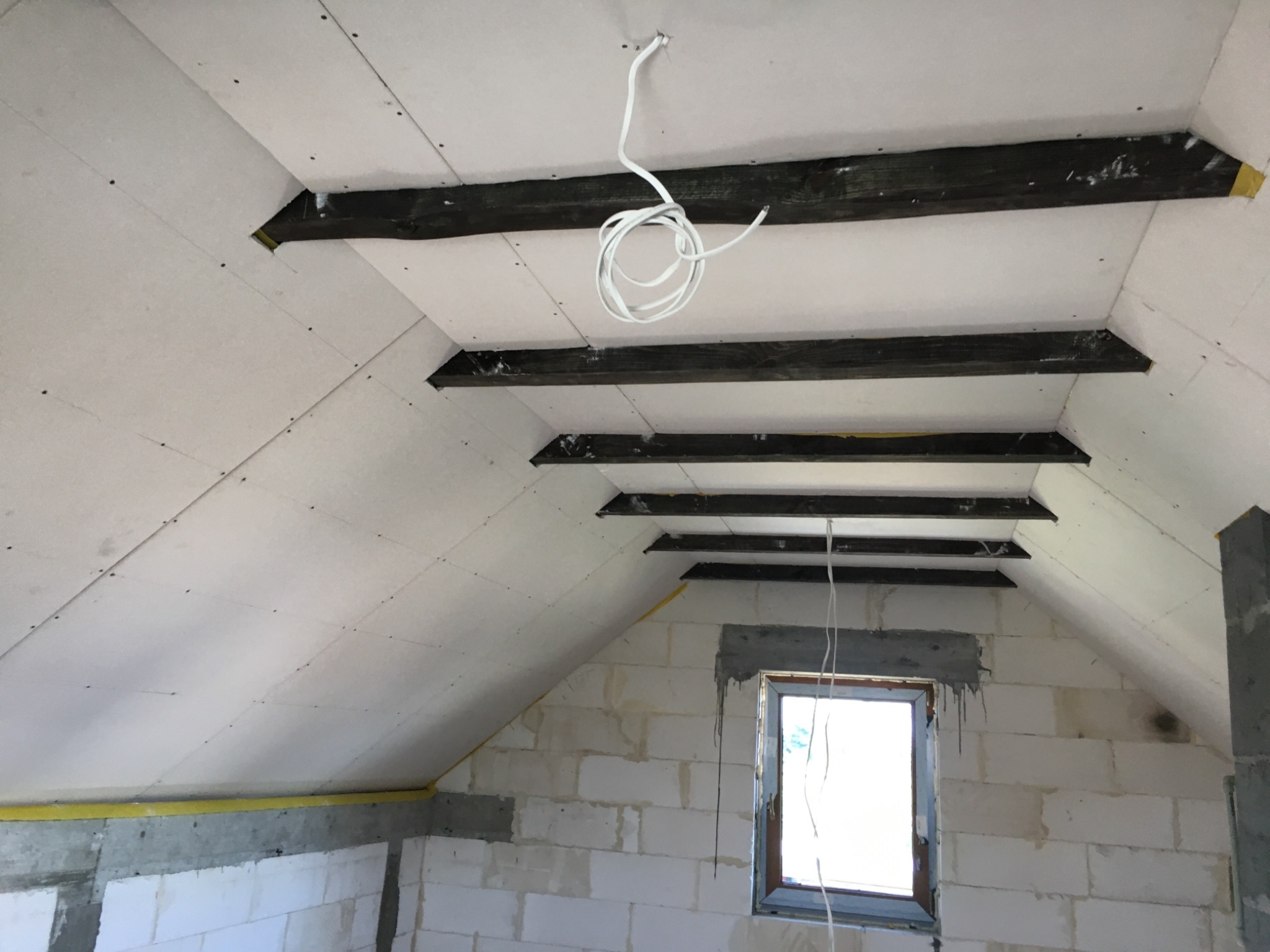 Poddasze w trakcie wykańczania: widoczne zamontowane płyty karton-gips na skośnym suficie z drewnianymi belkami, okno w surowej ścianie z białego bloczka, zwisający kabel elektryczny.