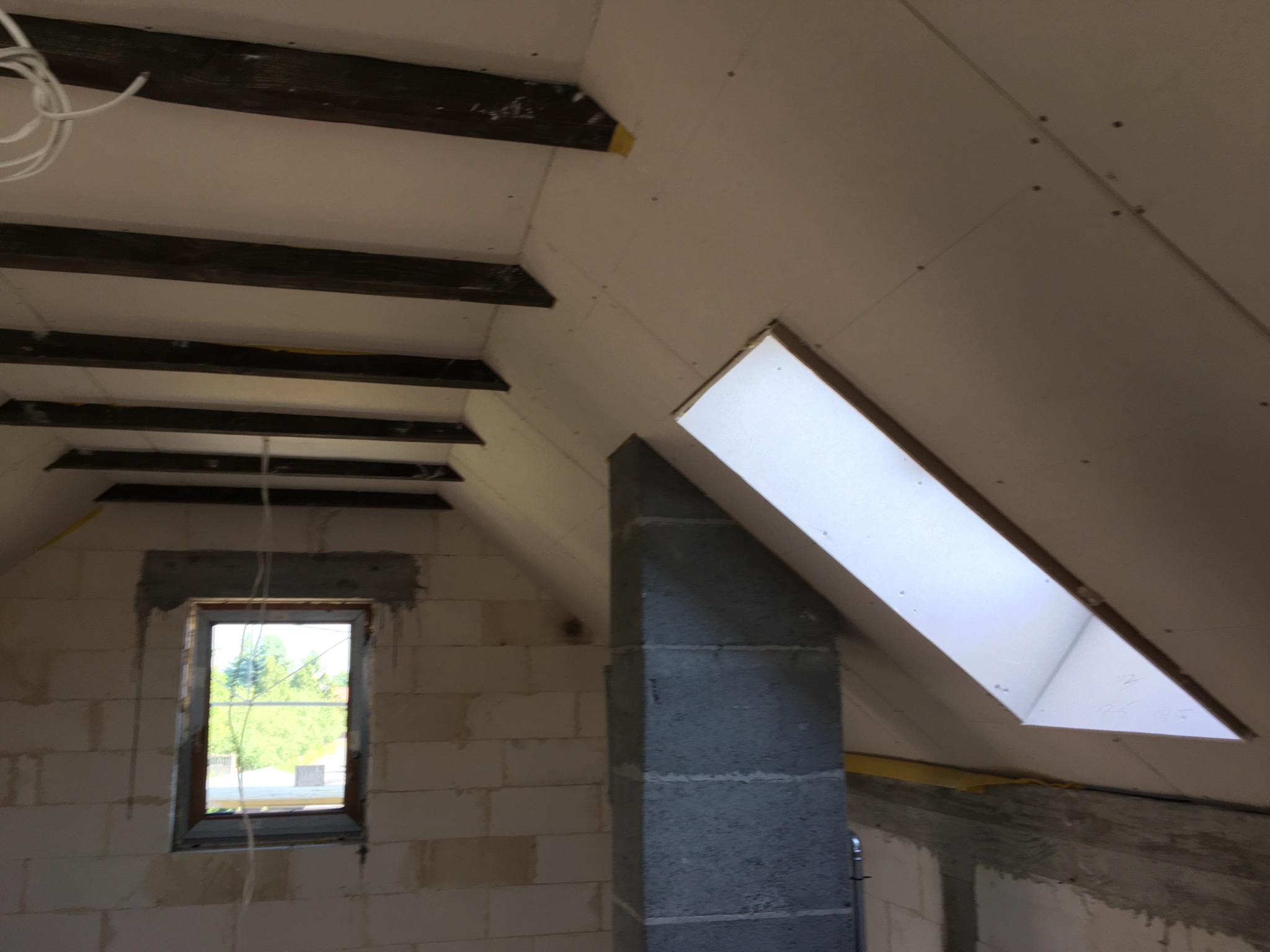 Poddasze w trakcie wykańczania: widoczne drewniane belki stropowe, okno dachowe, ściany z bloczków oraz zamontowane płyty kartonowo-gipsowe z widocznymi łączeniami.