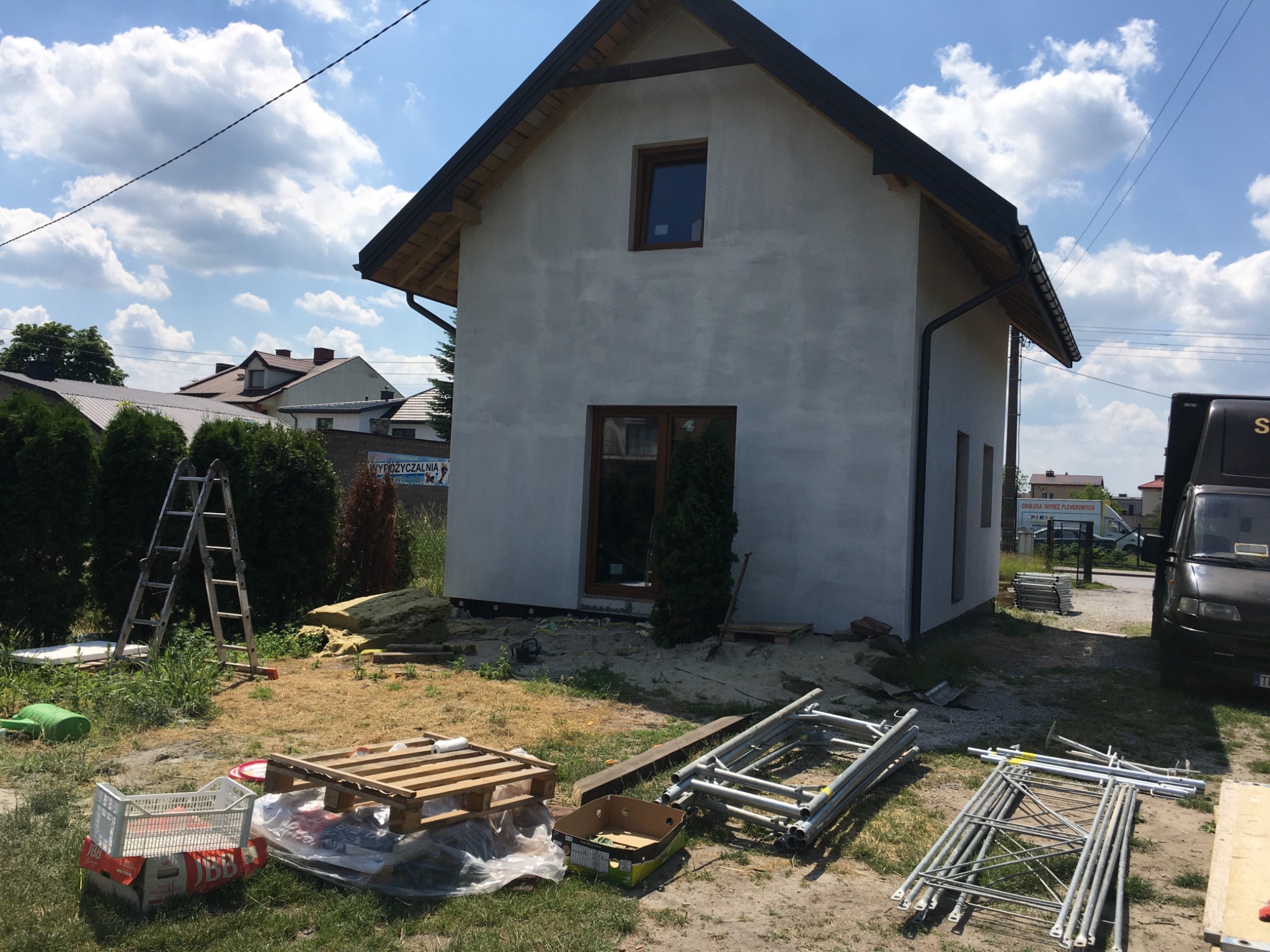 Dom jednorodzinny po nałożeniu szarego tynku, widoczne rusztowania, palety i materiały budowlane na trawniku, przygotowania do dalszych prac wykończeniowych elewacji.