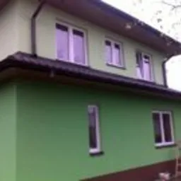 Dwukondygnacyjny dom z zieloną elewacją, brązowym cokołem i białymi oknami, widziany z boku, z drabinką opartą o ścianę.