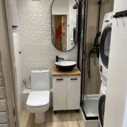 Nowoczesna łazienka z białą armaturą, czarną owalną umywalką na drewnianym blacie, kabiną prysznicową z czarnymi profilami i pralką w zabudowie.