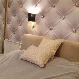 Wykończona sypialnia z tapicerowanym panelem ściennym w kolorze pudrowego różu, zdobionym kryształkami, podświetlonym listwą LED i kinkietem, z beżowym zagłówkiem i poduszkami.