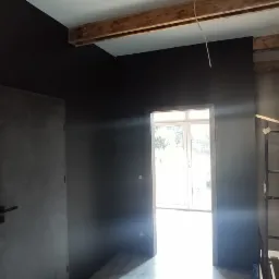 Wnętrze w trakcie remontu: ściany pomalowane na ciemny kolor, widoczne drewniane belki stropowe, drzwi i fragment rusztowania