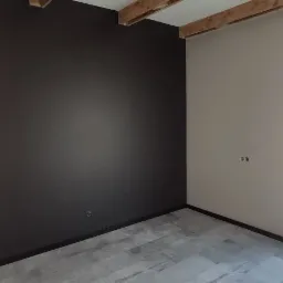 Pomieszczenie po częściowym odświeżeniu: jedna ściana pomalowana na grafitowo, druga w jasnym odcieniu, widoczne drewniane belki stropowe i szare panele podłogowe, puszki elektryczne na ścianie.