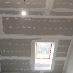 Poddasze w trakcie prac wykończeniowych: widoczne płyty kartonowo-gipsowe na skosach i suficie, szpachlowane łączenia i wkręty, okno dachowe, drabina przystawna i żarówka na kablu.