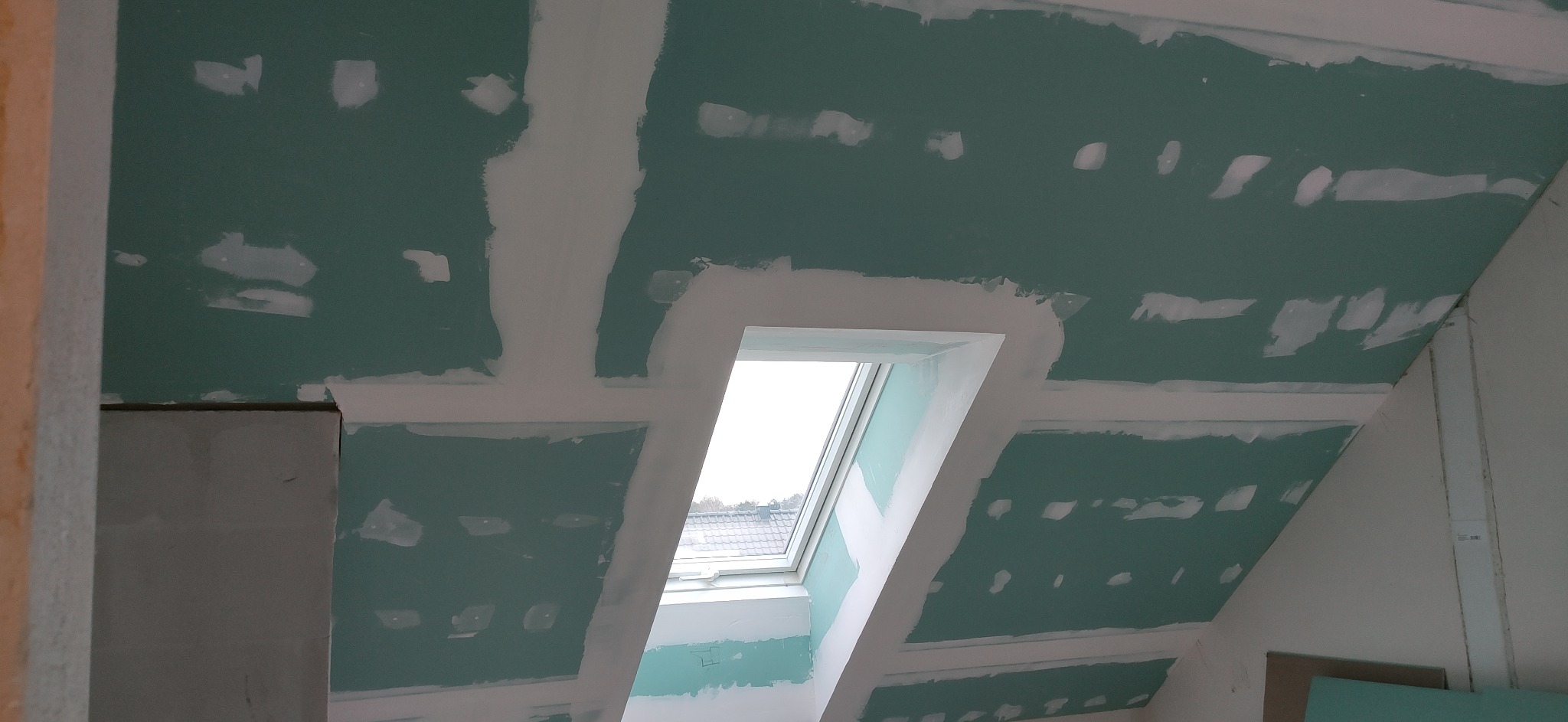 Poddasze z oknem dachowym w trakcie montażu płyt kartonowo-gipsowych; widoczne łączenia płyt pokryte masą szpachlową, surowe wykończenie, brak malowania.