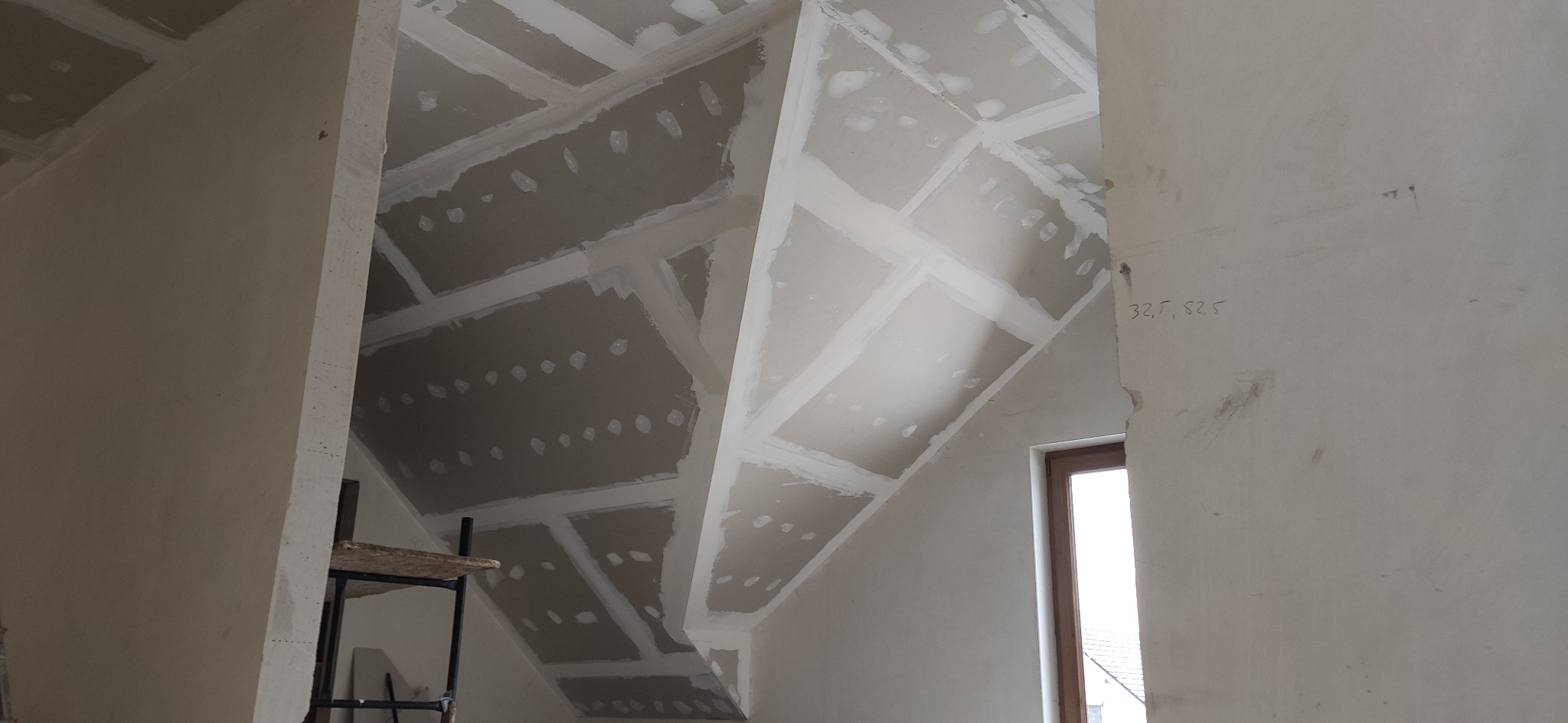 Skośny sufit na poddaszu z zamontowanymi płytami gipsowo-kartonowymi, widoczne łączenia płyt wypełnione masą szpachlową, fragment rusztowania i okno w tle.