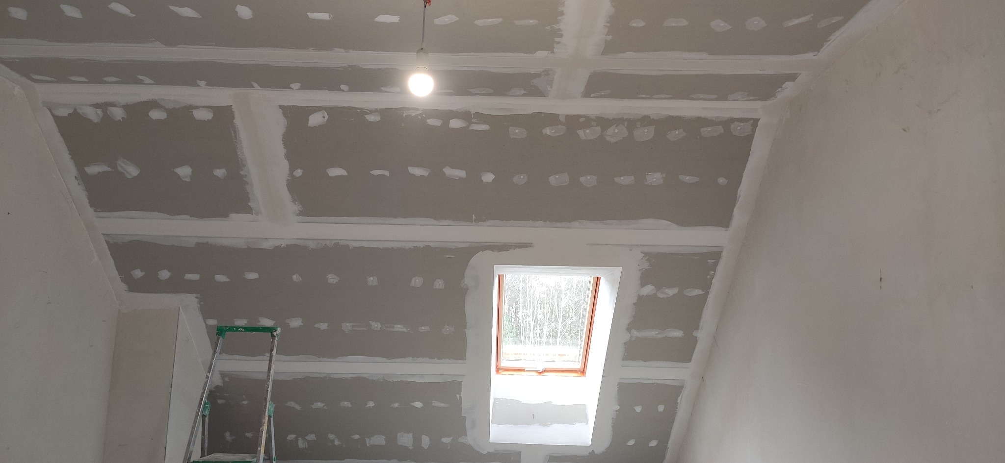 Poddasze w trakcie prac wykończeniowych: widoczne płyty kartonowo-gipsowe na skosach i suficie, szpachlowane łączenia i wkręty, okno dachowe, drabina przystawna i żarówka na kablu.