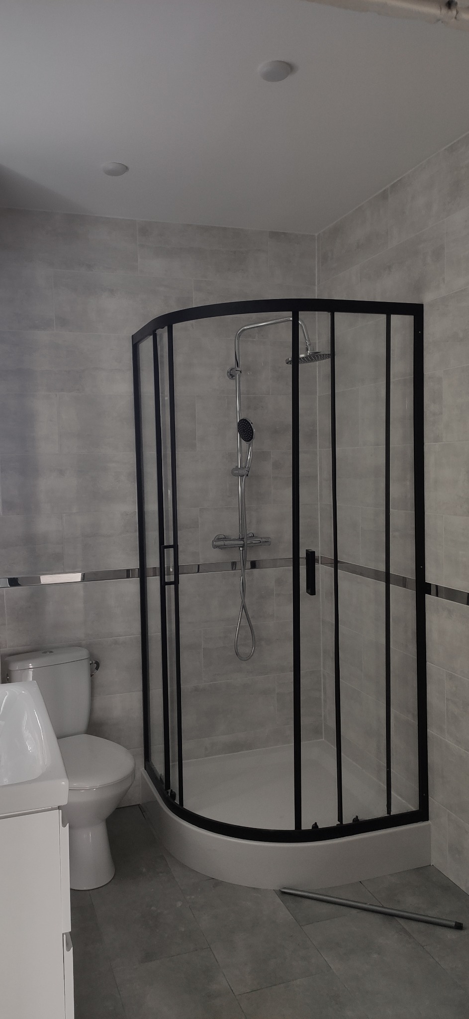 Wykończona łazienka z kabiną prysznicową z czarnymi ramami, szarymi płytkami i białą toaletą z umywalką; na podłodze leży metalowa rurka.