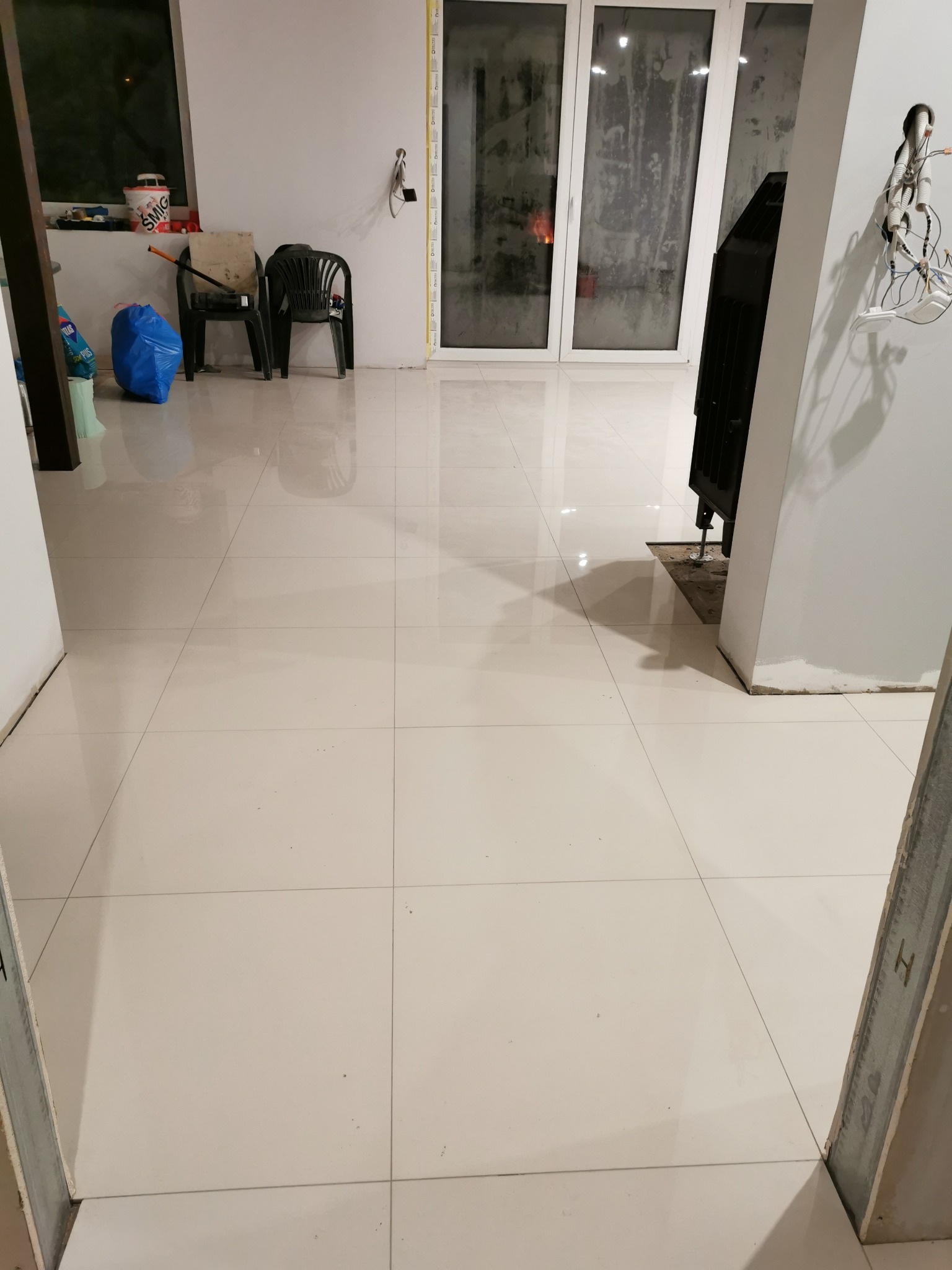 Białe, błyszczące płytki podłogowe w nowym, pustym pomieszczeniu z widocznym kominkiem, przygotowanym do montażu, z fragmentem ściany z wystającymi kablami i przewodami elektrycznymi, widok zza...