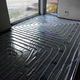 Rozłożona instalacja ogrzewania podłogowego z białych rur na czarnej macie z widocznym oknem i przygotowanym miejscem na ścianie pod termostat.