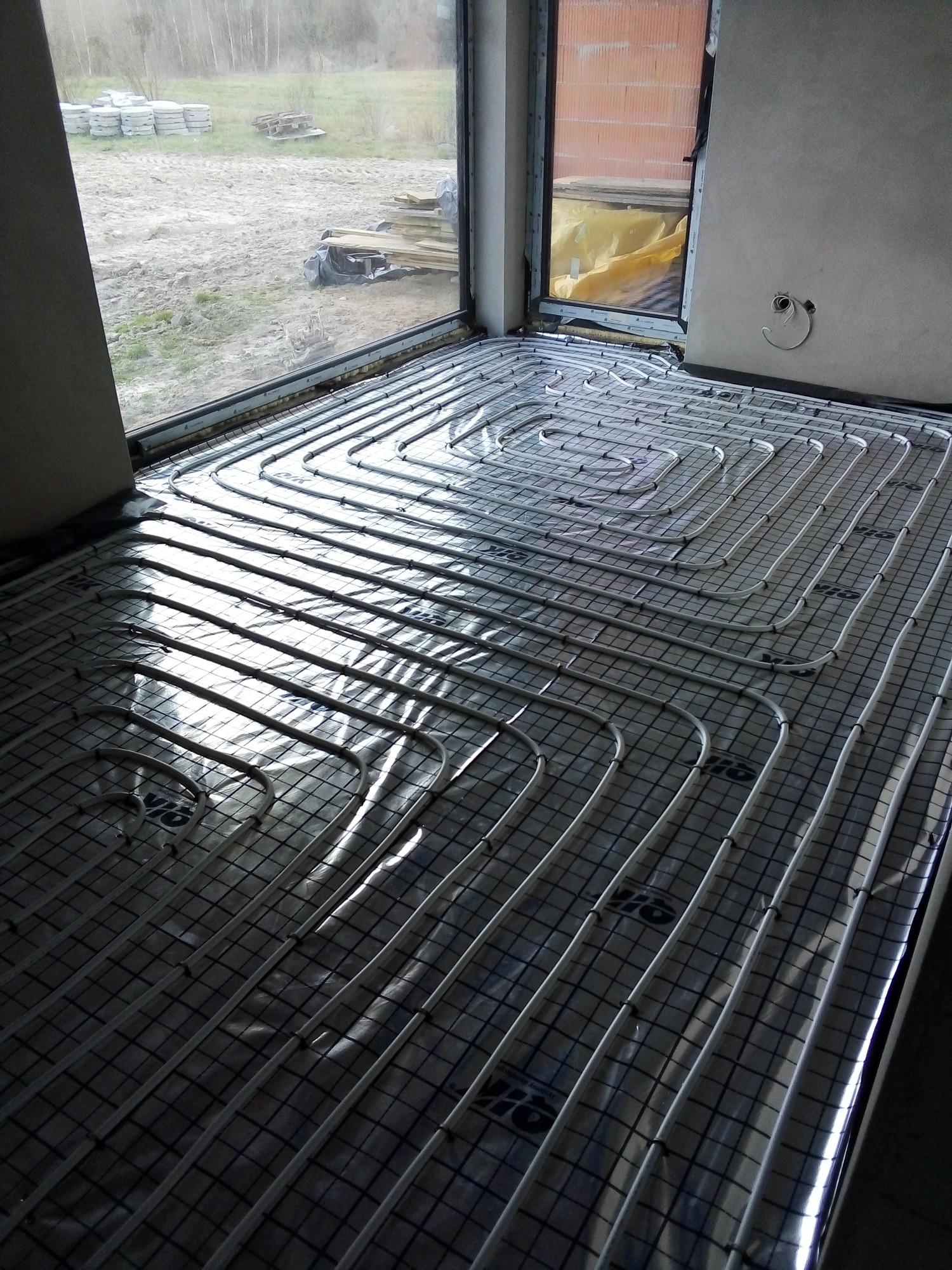 Rozłożona instalacja ogrzewania podłogowego z białych rur na czarnej macie z widocznym oknem i przygotowanym miejscem na ścianie pod termostat.