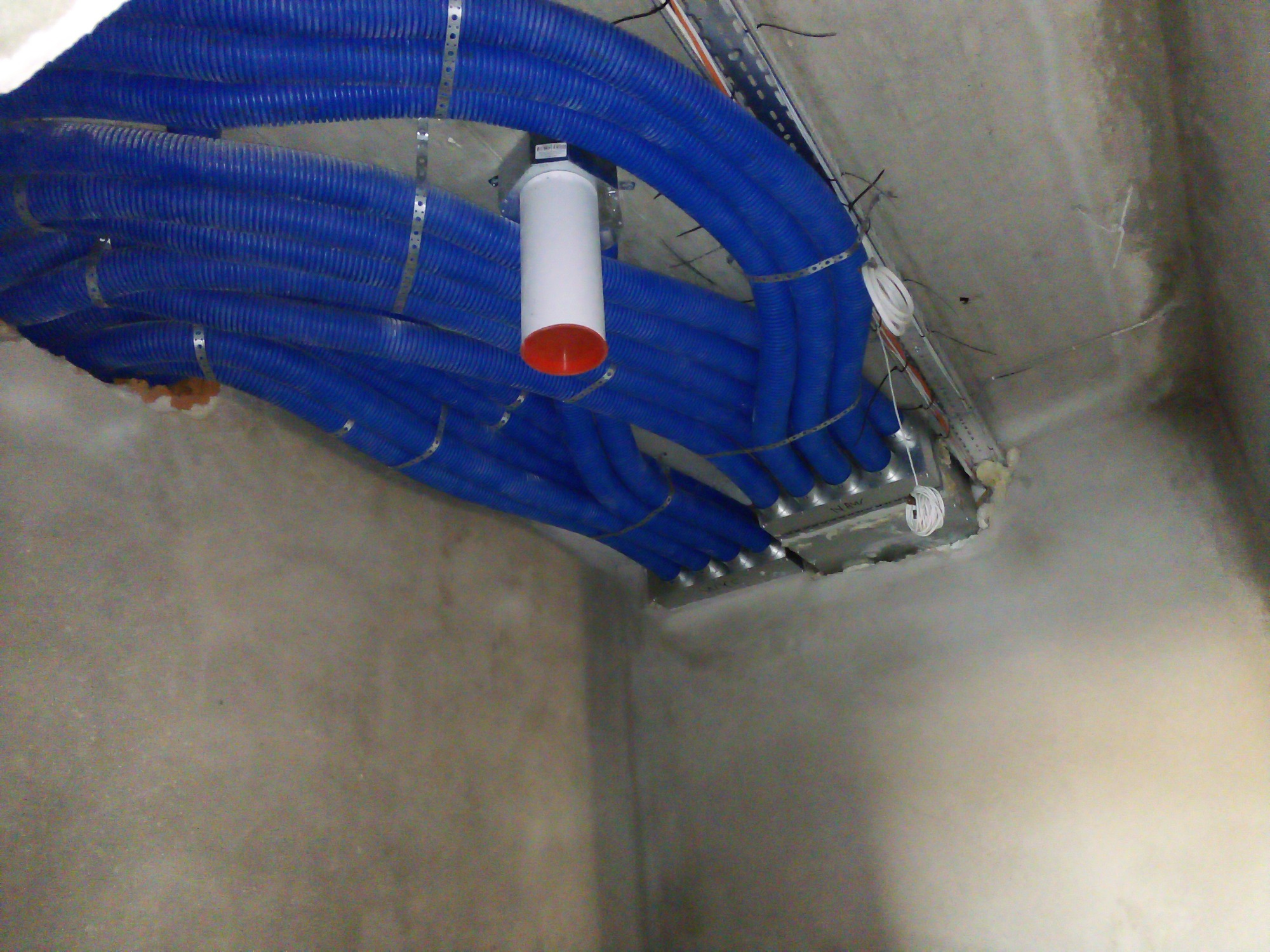 Niebieskie, karbowane rury wentylacyjne zamocowane do sufitu za pomocą metalowych opasek, z widocznym elementem instalacji w kolorze białym z czerwonym zakończeniem.