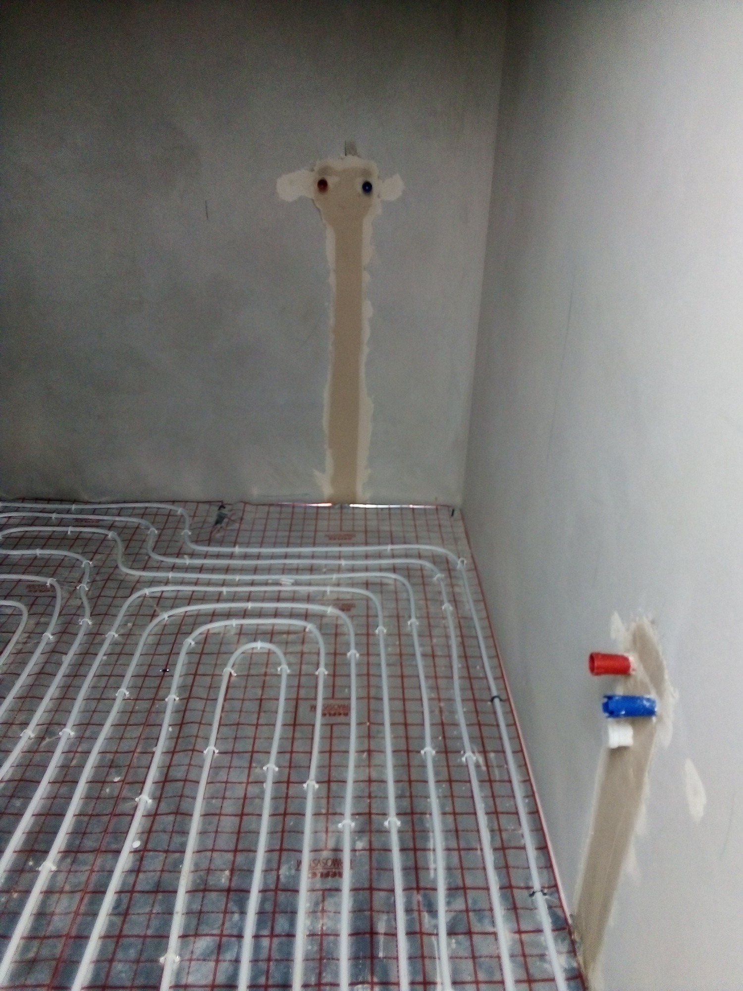 Montaż ogrzewania podłogowego: białe rurki ułożone na siatce na podłodze, z widocznymi wyprowadzeniami rur ze ściany z czerwonym i niebieskim korkiem.