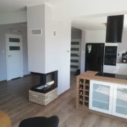 Nowoczesny salon z kominkiem narożnym obudowanym cegłą, widok na biało-czarną kuchnię z drewnianym barkiem i winami