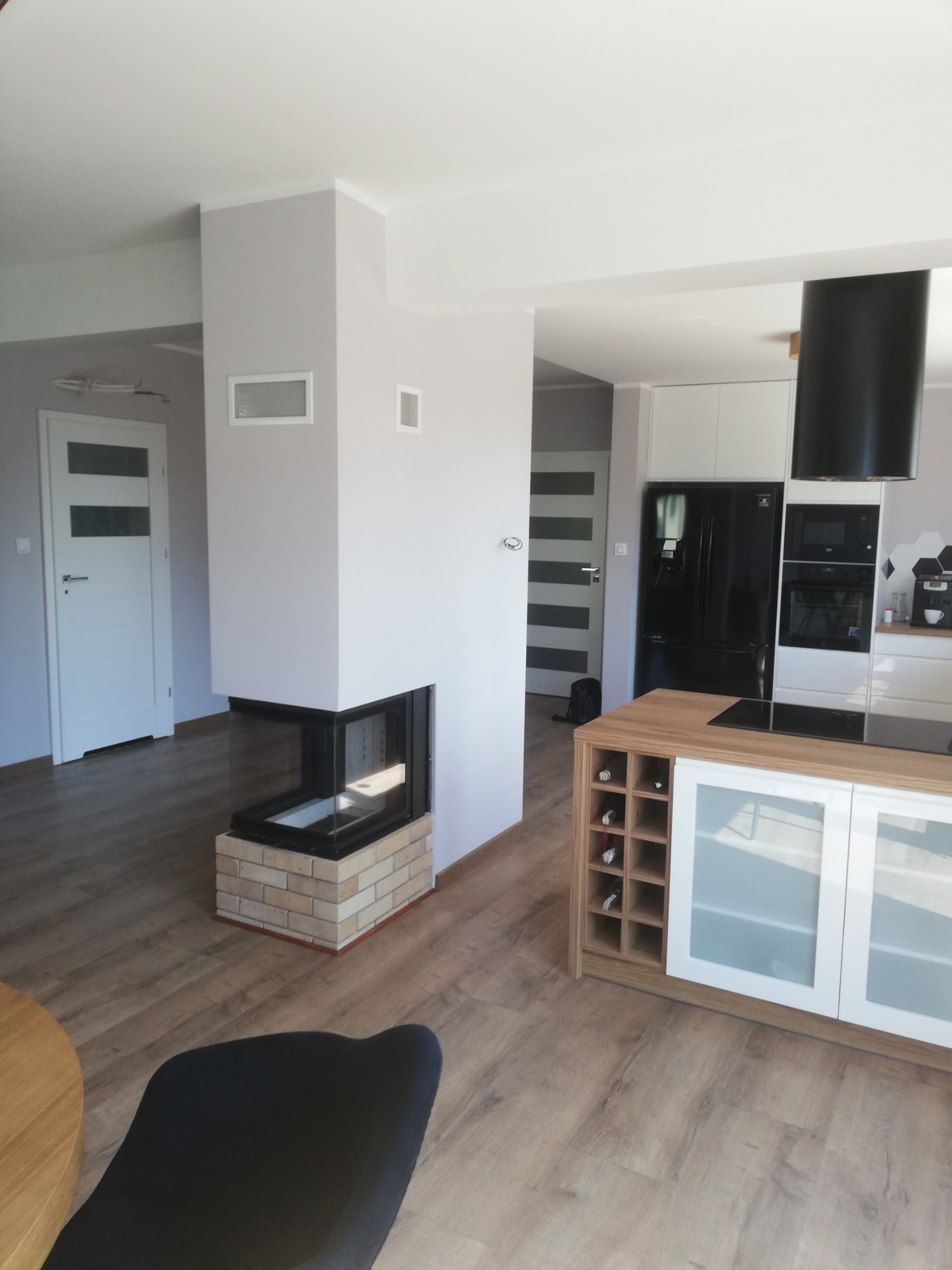 Nowoczesny salon z kominkiem narożnym obudowanym cegłą, widok na biało-czarną kuchnię z drewnianym barkiem i winami