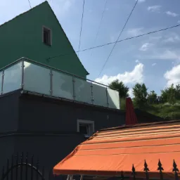 Szklana balustrada balkonowa z nierdzewnymi słupkami, zamontowana na ciemnoszarym balkonie przylegającym do zielonego domu, widok zza czarnego kutego ogrodzenia i pomarańczowej markizy.