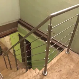 Wnętrze w trakcie remontu: drewniane schody z zamontowaną, prostą, metalową barierką ze stali nierdzewnej, widok z góry, w tle zielona ściana i sprzęt budowlany.