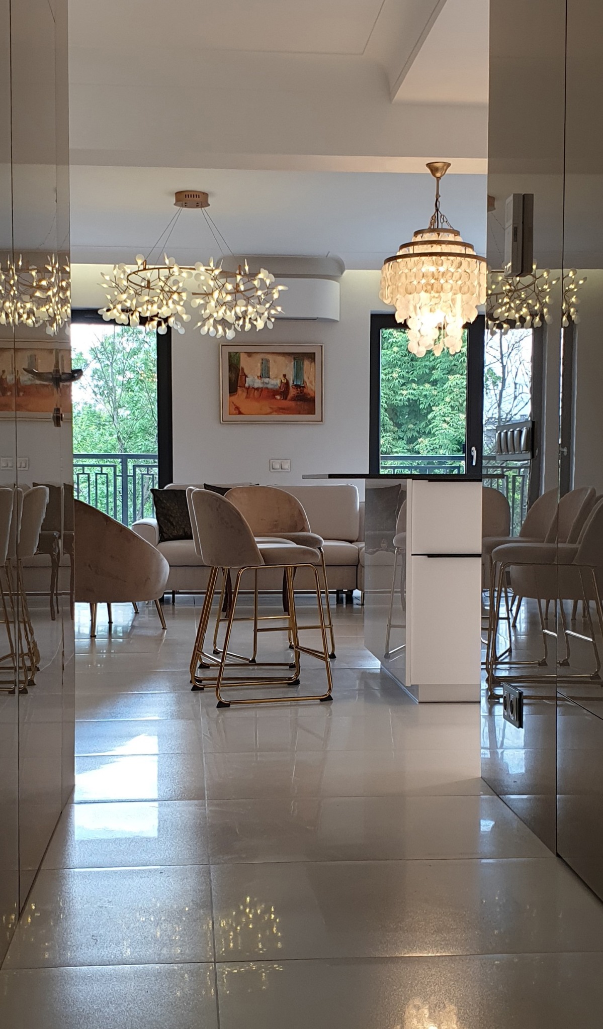 Salon w Kielcach widziany z korytarza, z widocznymi płytami akrylowymi na ścianach korytarza, błyszcząca podłoga, nowoczesne krzesła barowe z złotymi nogami, sofa w odcieniach beżu, okno z widokiem...