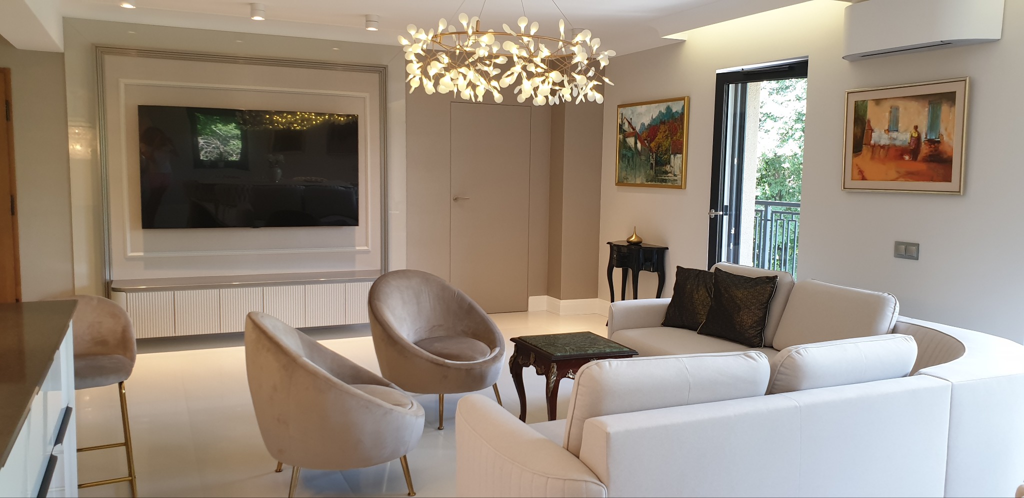 Jasny salon z białymi sofami, aksamitnymi fotelami na złotych nóżkach, telewizorem w ozdobnej ramie i nowoczesnym żyrandolem z motywem liści.