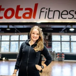 Uśmiechnięta blondynka w czarnej bluzie z kapturem, stojąca w siłowni z napisem 'total fitness' w tle, na nadgarstku widoczny sportowy zegarek.