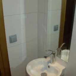 Mała łazienka z umywalką, baterią i lustrem, wykończona płytkami w odcieniach bieli i beżu z delikatnym wzorem poziomych linii, widoczny fragment drewnianej ościeżnicy drzwi.