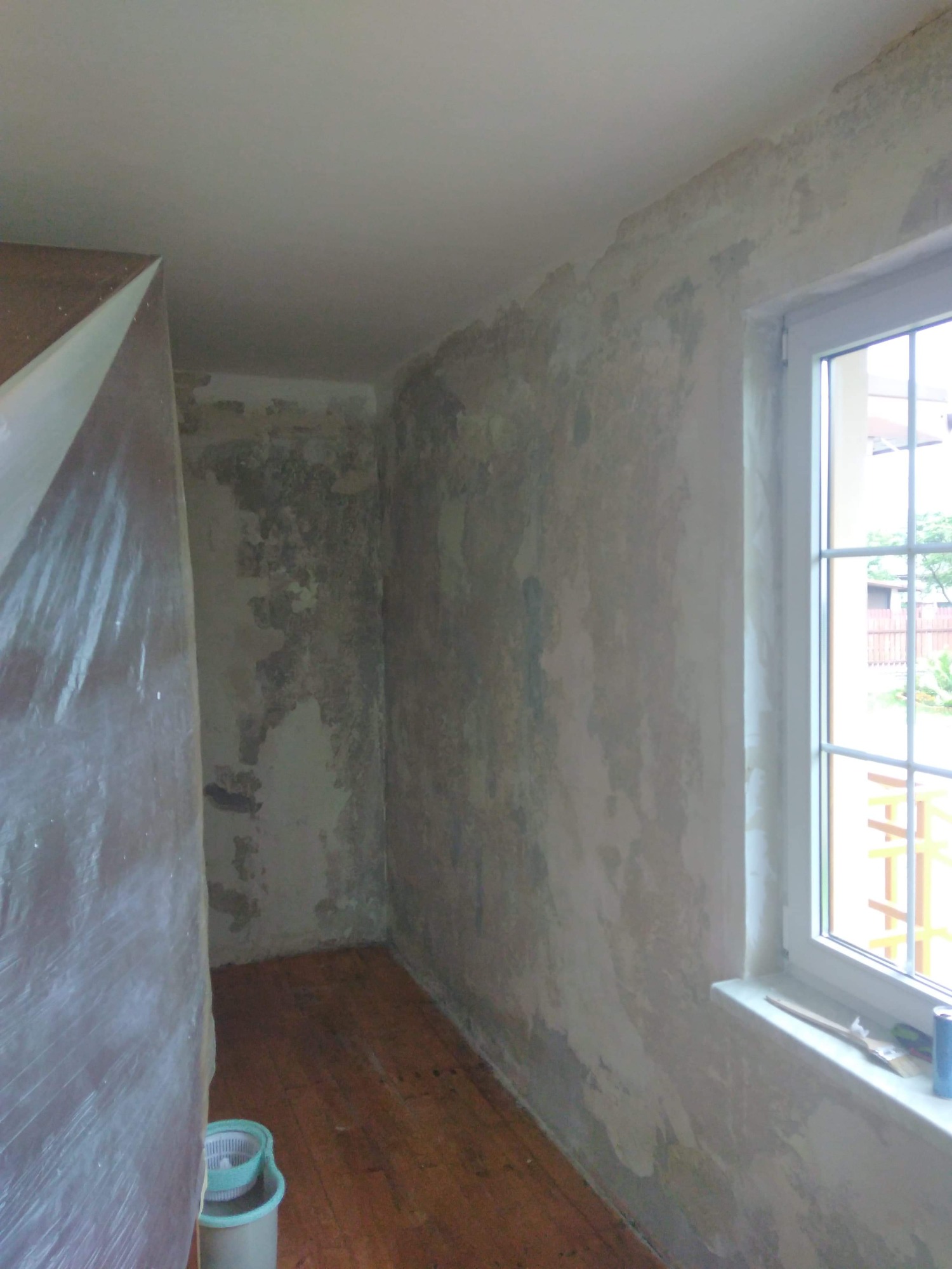 Pokój w trakcie przygotowania do malowania: ściany z nierównościami i pozostałościami starej farby, okno z pędzlem i puszką farby na parapecie, podłoga zabezpieczona, widoczny pojemnik na wodę.