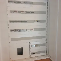 Otwarta, biała skrzynka rozdzielcza z bezpiecznikami i licznikami, zamontowana na surowej, ceglanej ścianie z widocznymi notatkami. Widoczne kable elektryczne.