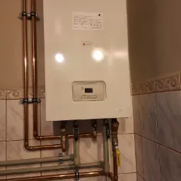 Zamontowany, biały piec gazowy marki Saunier Duval z widocznymi miedzianymi rurami, zaworami i elementami instalacji na tle ceramicznej ściany.