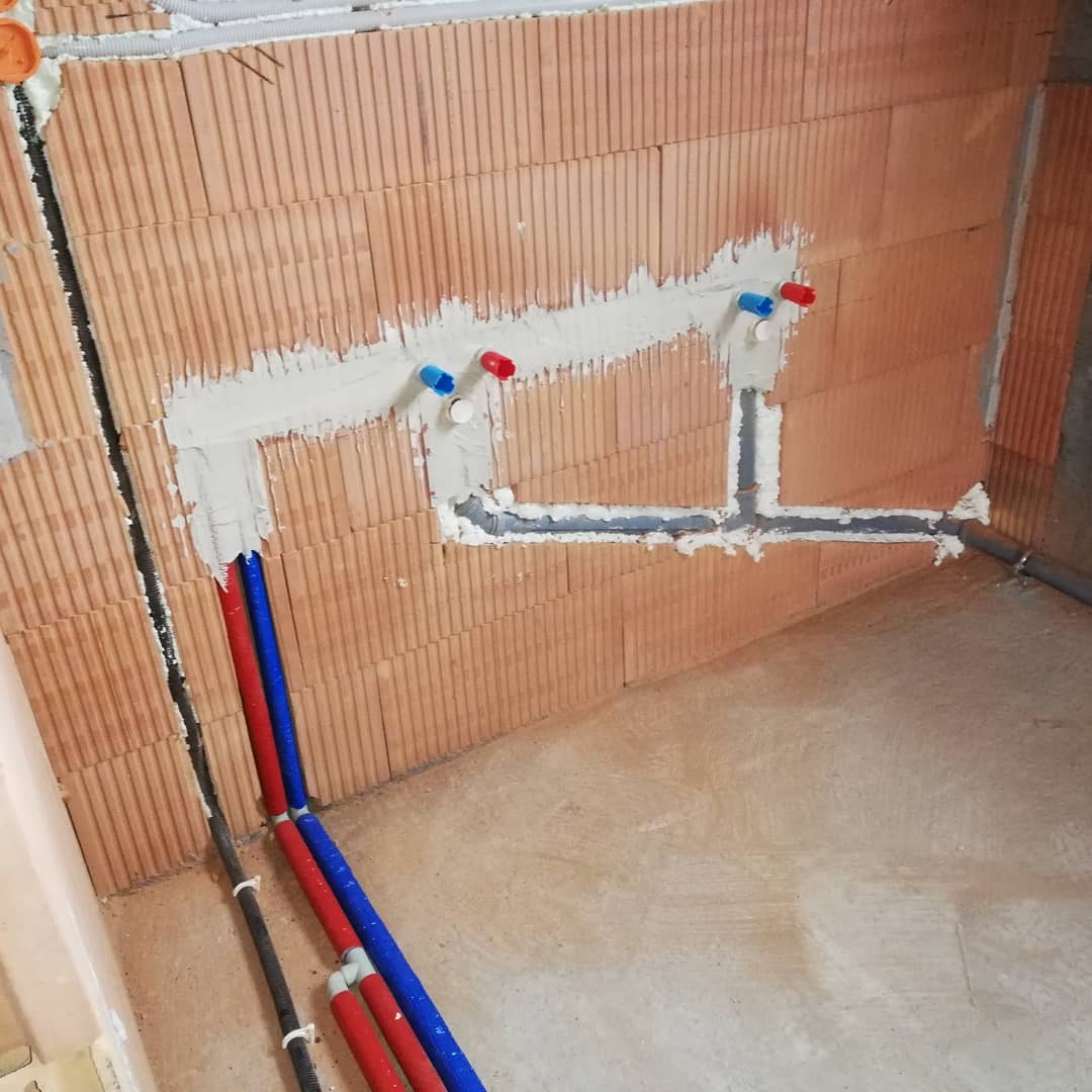 Instalacja wodna w trakcie montażu na ścianie z cegły ceramicznej, widoczne rury doprowadzające ciepłą i zimną wodę z czerwonymi i niebieskimi zaślepkami oraz szare rury odpływowe.