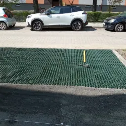 Instalacja zielonej kratki trawnikowej na przygotowanym podłożu, widoczna poziomica i gumowy młotek, w tle zaparkowane samochody.