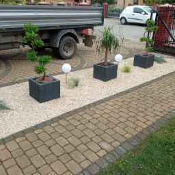 Aranżacja przestrzeni przed domem z użyciem trzech donic z drzewkami, białego żwiru, lamp ogrodowych i kostki brukowej, widoczny samochód dostawczy w tle.