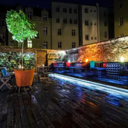 Oświetlony ogródek restauracyjny nocą w Katowicach, z drewnianym tarasem, ceglanymi ścianami, zieloną ścianą i podświetlanymi meblami.