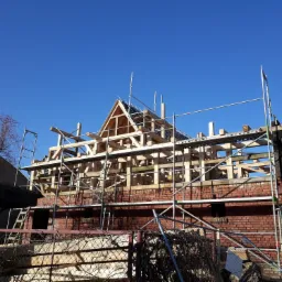 Budowa domu z czerwonej cegły w trakcie wznoszenia drewnianej konstrukcji dachu, widoczne rusztowania i błękitne niebo.