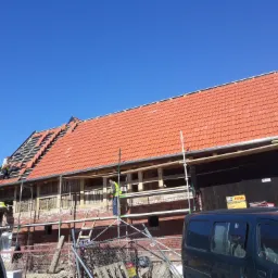 Prace dekarskie w Słupsku: ekipa montuje czerwoną dachówkę na konstrukcji więźby, widoczne rusztowania i częściowo ocieplona ściana budynku.