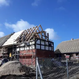 Budynek szkieletowy w trakcie renowacji dachu, widoczne rusztowania, drewniana konstrukcja więźby, częściowo pokryty dach deskami, w tle drugi budynek z nowym poszyciem dachowym, błękitne niebo...